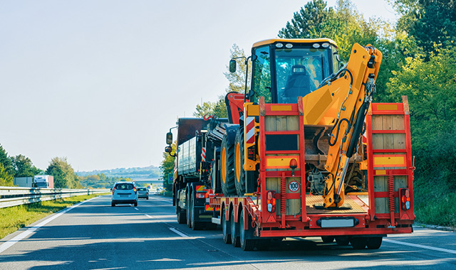 A heavy truck on a tree-lined highway pulls a trailer of large construction equipment     